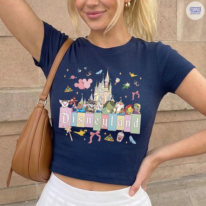 Pu&ograve; includere: Top corto blu navy con grafica colorata di Disneyland. Il design include un castello, vari personaggi dei cartoni animati e la scritta "Disneyland" in lettere pastello. La maglietta ha maniche corte e una silhouette aderente.