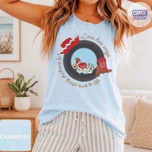 May include: A light blue sleeveless tank top featuring a graphic print of a black tire framing cartoon characters. The design includes a red cowboy hat, a red cowboy boot, and the text "Love has ways of bringing things back to life."