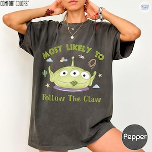 May include: A charcoal gray short-sleeve t-shirt featuring a graphic of a three-eyed green alien. The text reads "MOST LIKELY TO" above the alien and "Follow The Claw" below. Small icons of a rocket, blocks, a cactus, and stars surround the character.