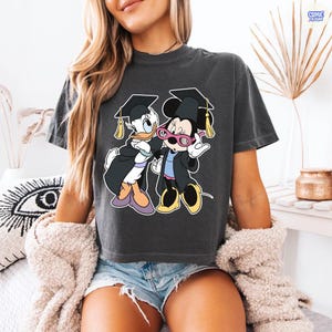 Puede incluir: Camiseta gris oscuro con un gr&aacute;fico de Pato Donald y Minnie Mouse con atuendo de graduaci&oacute;n. Pato Donald y Minnie Mouse llevan gorros y togas de graduaci&oacute;n. La camiseta tiene cuello redondo y mangas cortas.