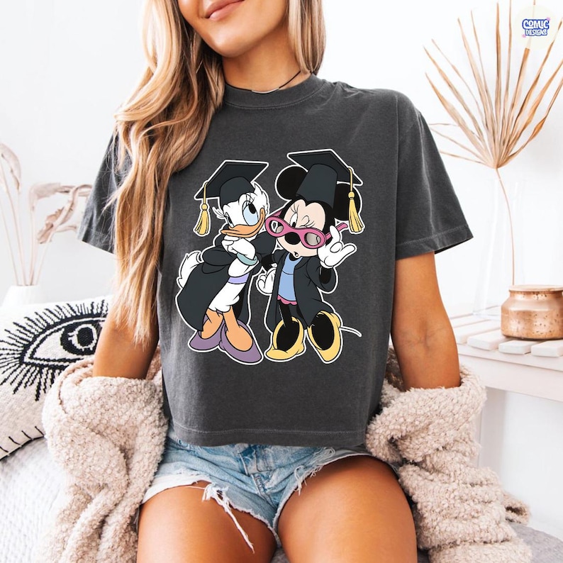 Puede incluir: Camiseta gris oscuro con un gr&aacute;fico de Pato Donald y Minnie Mouse con atuendo de graduaci&oacute;n. Pato Donald y Minnie Mouse llevan gorros y togas de graduaci&oacute;n. La camiseta tiene cuello redondo y mangas cortas.