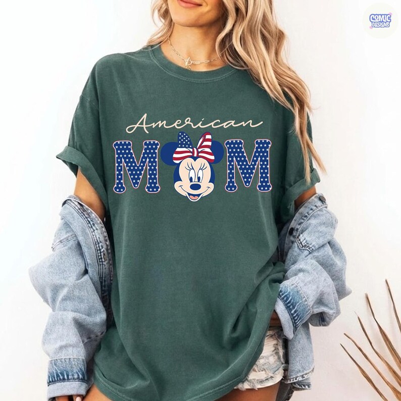 May include: A dark green t-shirt with the words "American MOM" in white script and blue block letters with a star pattern. The letter "O" is replaced with a Minnie Mouse graphic wearing a red, white, and blue bow.