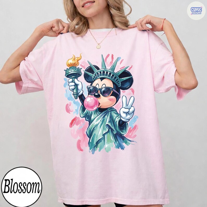 May include: A light pink t-shirt featuring a graphic of a cartoon mouse dressed as the Statue of Liberty, wearing sunglasses, blowing a pink bubble, holding a torch, and making a peace sign, with abstract pink and blue paint strokes in the background.