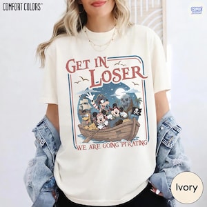 May include: Ivory-colored t-shirt with a pirate-themed graphic. The design features cartoon characters in a boat with the text "Get In Loser" and "We Are Going Pirating". The shirt has short sleeves and a crew neck.