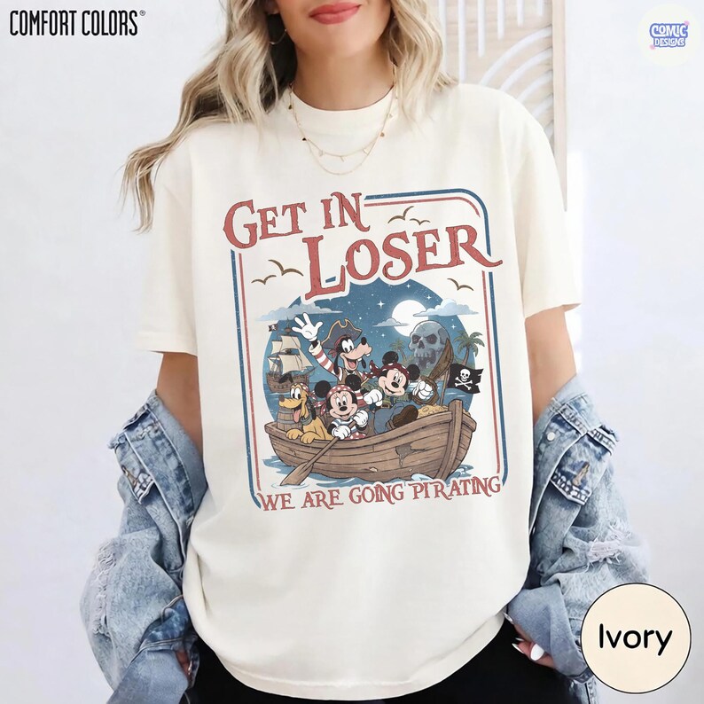 May include: Ivory-colored t-shirt with a pirate-themed graphic. The design features cartoon characters in a boat with the text "Get In Loser" and "We Are Going Pirating". The shirt has short sleeves and a crew neck.
