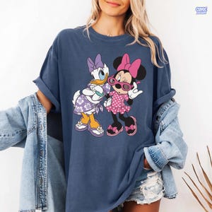 Puede incluir: Una camiseta de manga corta azul oscuro con un estampado gr&aacute;fico de Daisy Duck y Minnie Mouse. Daisy lleva un vestido y un lazo con flores moradas, mientras que Minnie lleva un vestido de lunares rosa, un lazo rosa y gafas de sol rosas.