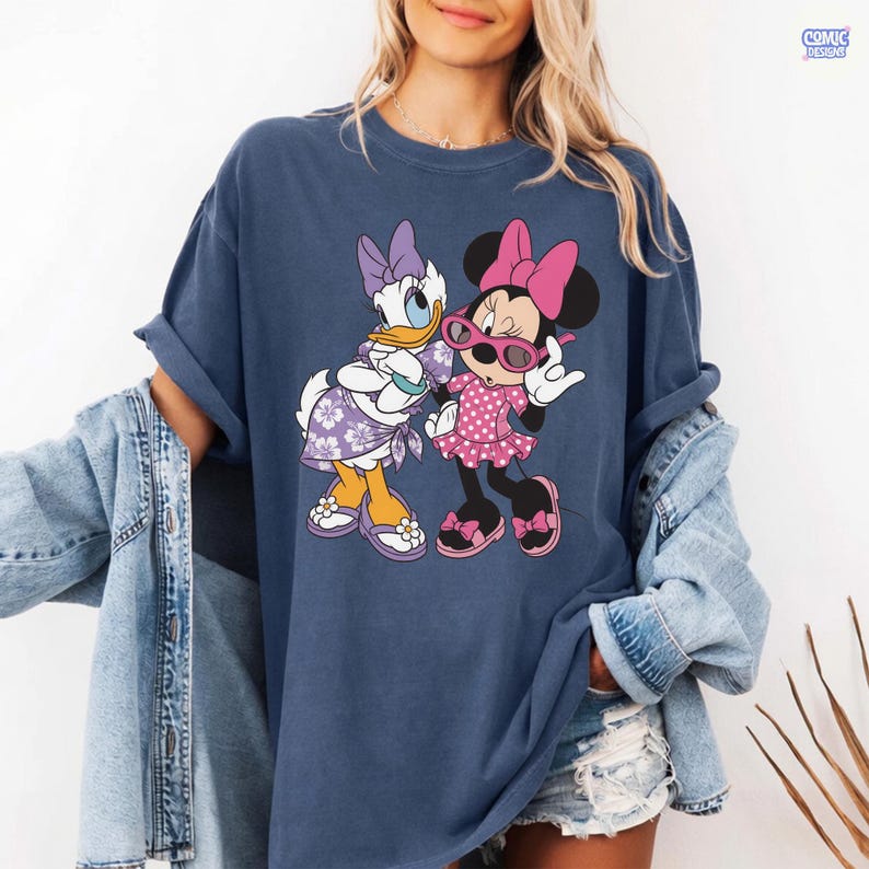 Puede incluir: Una camiseta de manga corta azul oscuro con un estampado gr&aacute;fico de Daisy Duck y Minnie Mouse. Daisy lleva un vestido y un lazo con flores moradas, mientras que Minnie lleva un vestido de lunares rosa, un lazo rosa y gafas de sol rosas.