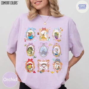 Puede incluir: Camiseta color orqu&iacute;dea con una colecci&oacute;n de retratos enmarcados de personajes de dibujos animados. Los retratos est&aacute;n en marcos ornamentados con lazos y detalles florales. La camiseta tiene los logotipos "COMFORT COLORS" y "COMIC DESIGNS".