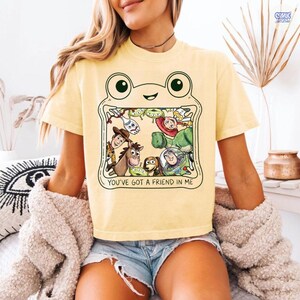 May include: A pale yellow short-sleeve t-shirt featuring a graphic of a frog outline framing various animated characters from the Toy Story franchise. The text below the characters reads, "YOU'VE GOT A FRIEND IN ME" in a simple black font.