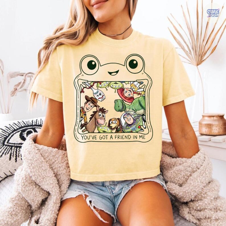 May include: A pale yellow short-sleeve t-shirt featuring a graphic of a frog outline framing various animated characters from the Toy Story franchise. The text below the characters reads, "YOU'VE GOT A FRIEND IN ME" in a simple black font.