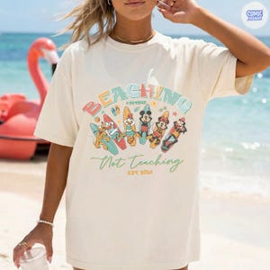 May include: Cream-colored t-shirt with the words "BEACHING #Retired Not Teaching EST. 2026" in a colorful design. The shirt features cartoon characters on surfboards, with a beach and ocean background.