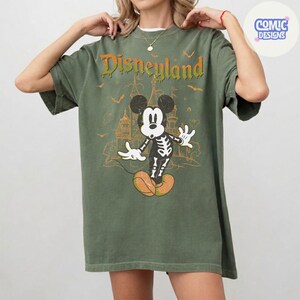 Może przedstawiać: Oliwkowozielony t-shirt z kr&oacute;tkim rękawem z grafiką szkieletu Myszki Miki na tle zarysu zamku. Napis "Disneyland" widnieje w ociekającej pomarańczowej czcionce nad postacią, w otoczeniu małych nietoperzy i gwiazdek.