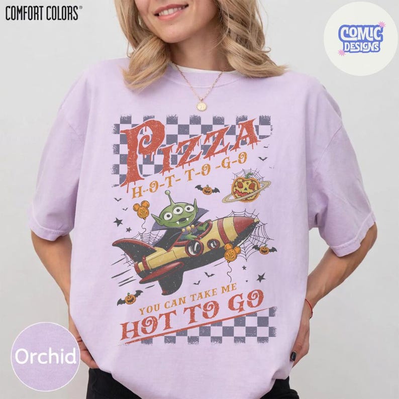 May include: A light purple t-shirt featuring a graphic of a green three-eyed alien in a yellow and red rocket ship. The text reads Pizza H-O-T-T-O-G-O and You can take me HOT TO GO, surrounded by spider webs, bats, and pumpkin illustrations.