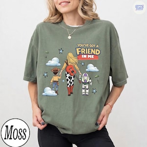 May include: A moss green crewneck t-shirt featuring a graphic of three characters walking away, with the text "YOU'VE GOT A FRIEND IN ME". The design includes a cowboy, a cowgirl with a guitar, and a space ranger, surrounded by clouds and stars.