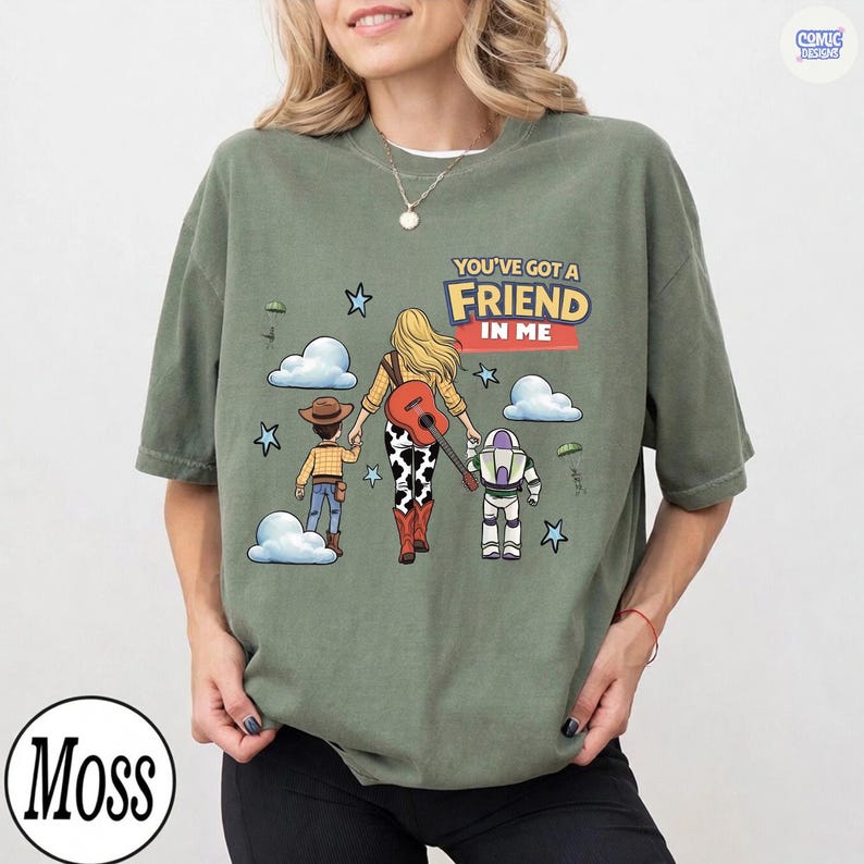 May include: A moss green crewneck t-shirt featuring a graphic of three characters walking away, with the text "YOU'VE GOT A FRIEND IN ME". The design includes a cowboy, a cowgirl with a guitar, and a space ranger, surrounded by clouds and stars.