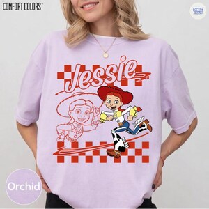 May include: A light purple short-sleeve t-shirt featuring a graphic of the character Jessie from Toy Story. The design includes the name Jessie in script, a red and white checkered pattern, and a colorful illustration of the character.
