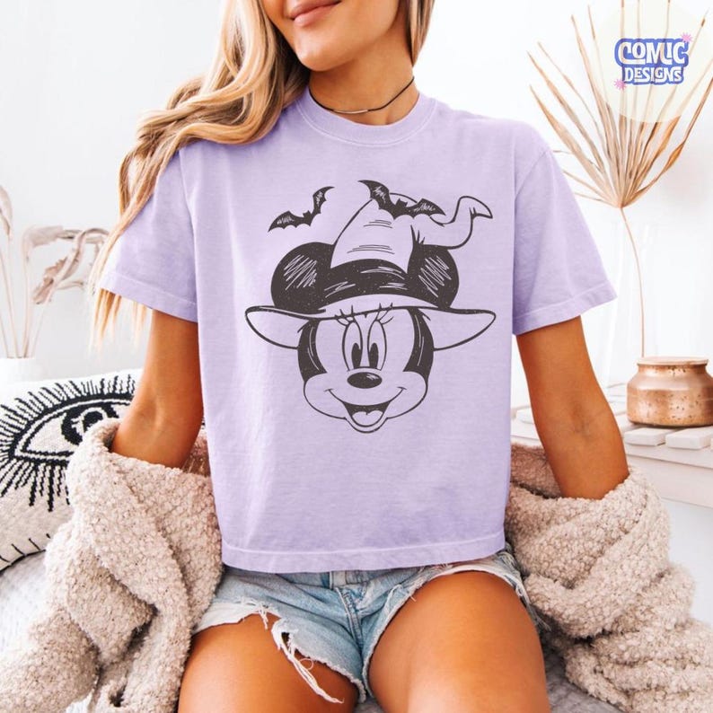 May include: A light purple t-shirt featuring a black and white graphic of Minnie Mouse wearing a witch hat with bats. The shirt has short sleeves and a relaxed fit. The text "COMIC DESIGNS" is in the upper right corner.