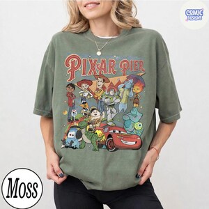 Pu&ograve; includere: T-shirt verde oliva con una grafica colorata di personaggi Pixar e la scritta "Pixar Pier". Il design include personaggi di Toy Story, Cars e altri film Pixar. La maglietta ha un girocollo e maniche corte.