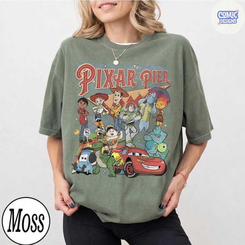 Pu&ograve; includere: T-shirt verde oliva con una grafica colorata di personaggi Pixar e la scritta "Pixar Pier". Il design include personaggi di Toy Story, Cars e altri film Pixar. La maglietta ha un girocollo e maniche corte.