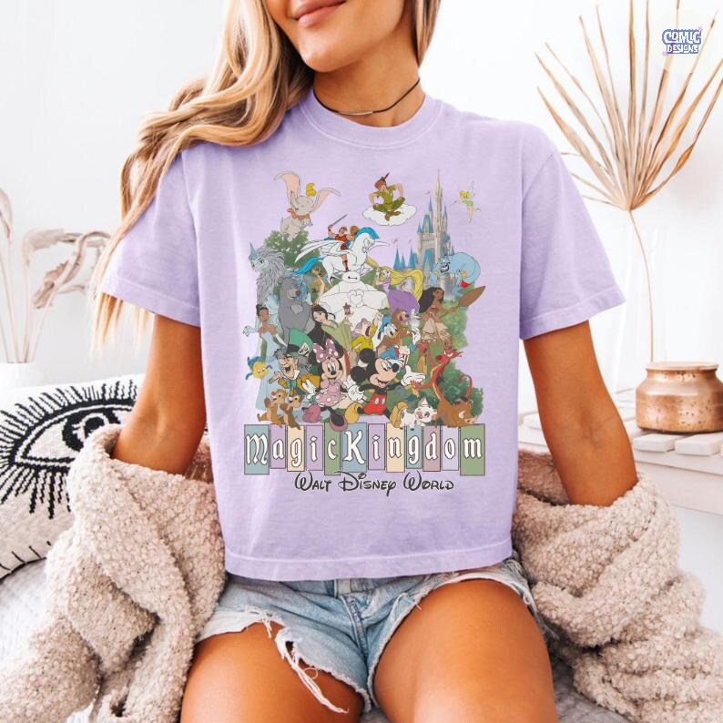 Puede incluir: Camiseta lila claro con un gr&aacute;fico colorido de personajes de Disney, incluyendo Mickey Mouse, Dumbo y Peter Pan. El texto "Magic Kingdom Walt Disney World" se muestra debajo de los personajes.