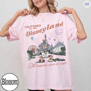 Op de afbeelding: Lichtroze Disneyland T-shirt met een vintage grafisch ontwerp. Het ontwerp bevat het Disneyland kasteel, Mickey en Minnie Mouse, en de tekst "California Disneyland Est. 1955". Het shirt heeft ook de tekst "The happiest place on earth".