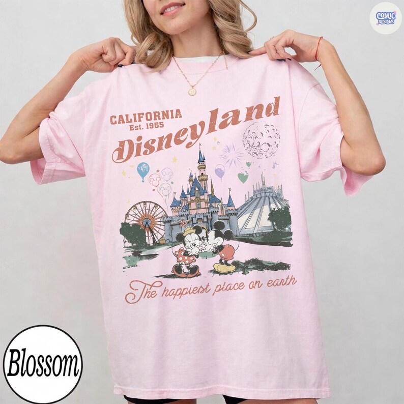 Op de afbeelding: Lichtroze Disneyland T-shirt met een vintage grafisch ontwerp. Het ontwerp bevat het Disneyland kasteel, Mickey en Minnie Mouse, en de tekst "California Disneyland Est. 1955". Het shirt heeft ook de tekst "The happiest place on earth".