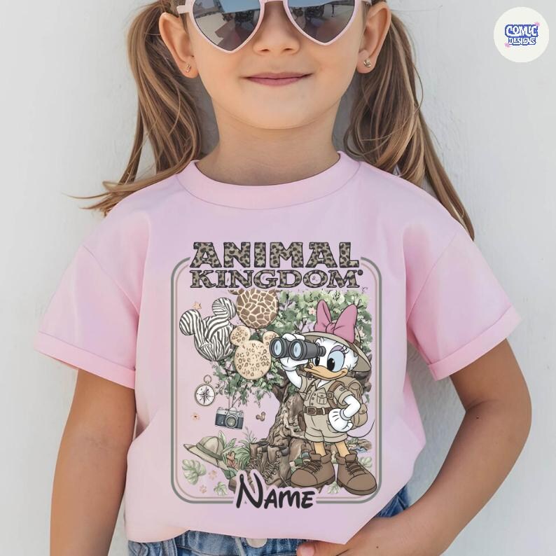 May include: A light pink t-shirt featuring a cartoon graphic with the text "ANIMAL KINGDOM". The graphic includes Daisy Duck with binoculars, balloons, and a tree with animal print patterns. The word "Name" is also visible.