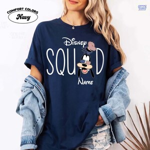 May include: Navy blue t-shirt with the words "Disney Squad" in white. The letter "D" features a cartoon character with an American flag-themed hat and sunglasses. The word "Name" is below the character. The shirt has the words "Comfort Colors" above the shirt color.
