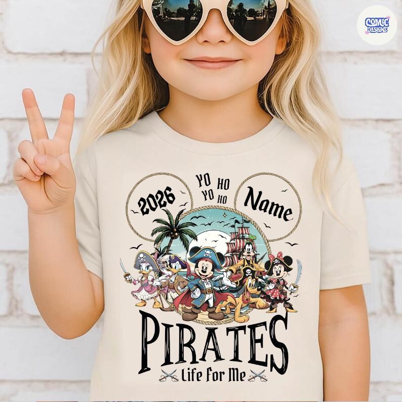 May include: A beige t-shirt featuring a pirate-themed graphic with Mickey Mouse and friends. The design includes the text "2026", "Yo Ho Ho", "Name", "Pirates", and "Life for Me". The child is wearing heart-shaped sunglasses and making a peace sign.