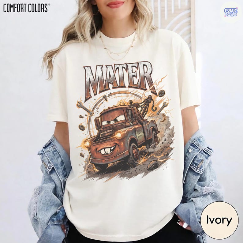 Pode incluir: Camiseta cor marfim com uma imagem de desenho animado do Mate do filme Carros. O gr&aacute;fico inclui o nome "MATER" em uma fonte desgastada acima do personagem, com ferramentas e raios ao redor do caminh&atilde;o. A camiseta &eacute; rotulada "COMFORT COLORS".