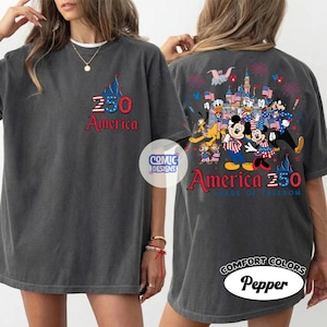 以下が含まれることがあります： 前面に城のグラフィックと赤と青の「250 America」の文字が入った濃い灰色のTシャツ。背面には、花火と旗を持つディズニーキャラクターのカラフルなイラストと、「America 250 Years of Freedom」の文字が描かれています。