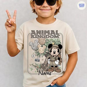 May include: Beige t-shirt featuring a graphic of Mickey Mouse in safari attire, with the text "ANIMAL KINGDOM" and a space for a name. The design includes Mickey Mouse-shaped balloons with animal print patterns and safari-themed elements.