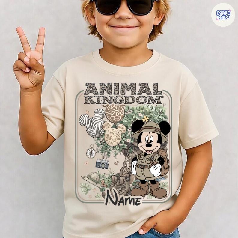 May include: Beige t-shirt featuring a graphic of Mickey Mouse in safari attire, with the text "ANIMAL KINGDOM" and a space for a name. The design includes Mickey Mouse-shaped balloons with animal print patterns and safari-themed elements.
