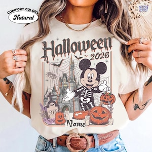 May include: Natural-coloured t-shirt with a Halloween 2026 graphic. The design features Mickey Mouse in a skeleton costume, a haunted castle, pumpkins, bats, and spiderwebs. The shirt also has the text "Name" at the bottom.