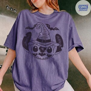 May include: A purple t-shirt featuring a black graphic of a cartoon character wearing a witch hat, with two bats above. The design is centered on the shirt. The shirt has short sleeves and a crew neck.