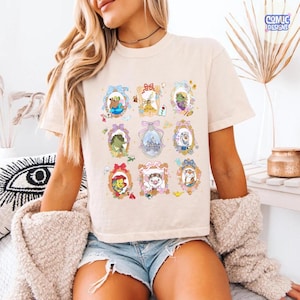 Puede incluir: Camiseta color crema con nueve retratos enmarcados de personajes de dibujos animados. Cada retrato tiene un marco ornamental &uacute;nico con colores pastel. La camiseta tiene mangas cortas y un corte holgado. El logotipo "Comic Designs" es visible en la esquina superior derecha.