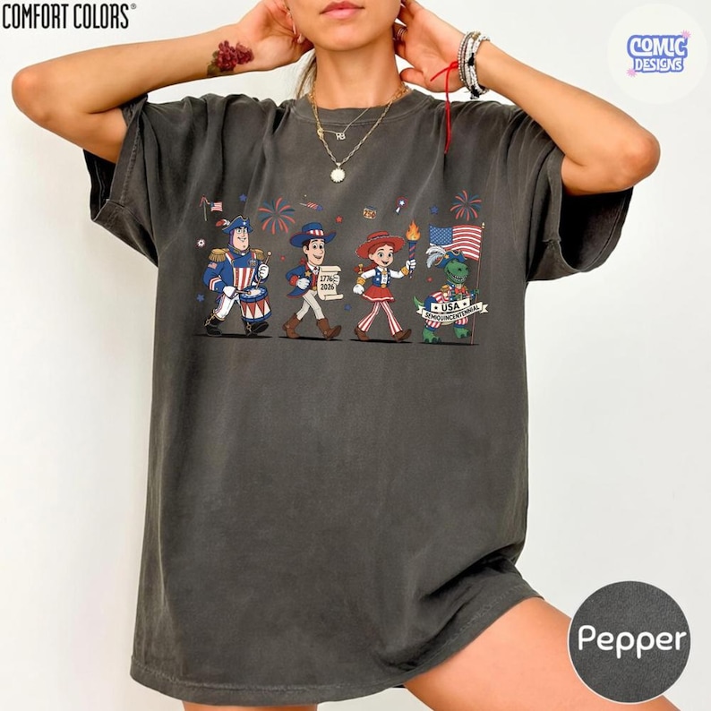 May include: Dark grey Comfort Colors t-shirt featuring a cartoon parade with characters in patriotic attire. The design includes fireworks, flags, and the text "USA Bicentennial". The shirt is a Pepper color.
