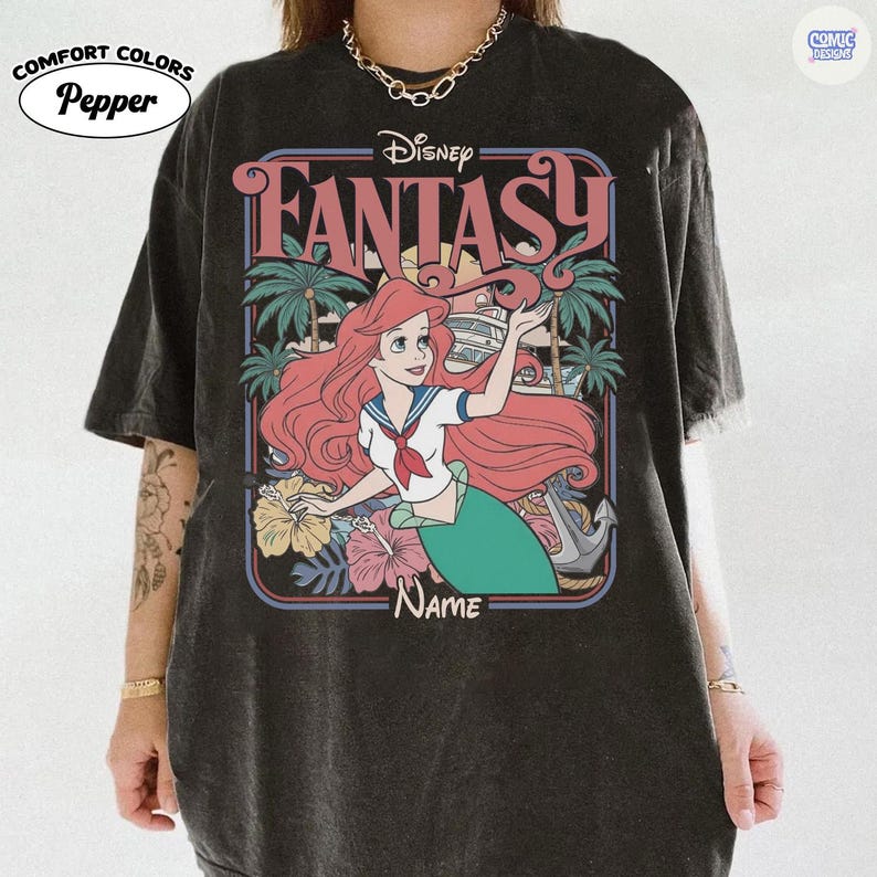 Op de afbeelding: Zwart Comfort Colors T-shirt met een Disney "Fantasy" ontwerp. De afbeelding bevat Ariel, palmbomen en tropische bloemen. Het woord "Name" staat onder de illustratie. Het shirt heeft ook het "Comic Designs" logo.