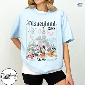 Op de afbeelding: Lichtblauw T-shirt met een Disneyland 2026-ontwerp met vuurwerk en Disney-figuren. Het ontwerp bevat Mickey en Minnie Mouse, Donald Duck en Goofy. Het shirt heeft een ronde hals en korte mouwen.