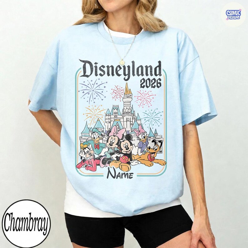 Op de afbeelding: Lichtblauw T-shirt met een Disneyland 2026-ontwerp met vuurwerk en Disney-figuren. Het ontwerp bevat Mickey en Minnie Mouse, Donald Duck en Goofy. Het shirt heeft een ronde hals en korte mouwen.