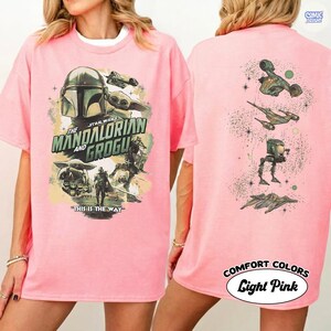 Op de afbeelding: Lichtroze T-shirt met een Star Wars-ontwerp met The Mandalorian en Grogu. De voorkant toont personages en tekst, terwijl de achterkant verschillende ruimtevaartuigen toont. Het shirt is gelabeld "COMFORT COLORS Light Pink."
