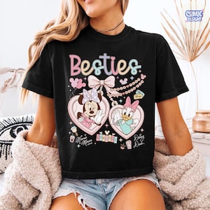 May include: Black t-shirt featuring the word "Besties" in pastel colors. The shirt showcases Minnie Mouse and Daisy Duck in heart-shaped lockets. Additional graphics include balloons, bows, and musical notes. The shirt is a comfortable, casual style.