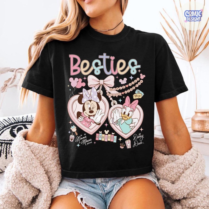 May include: Black t-shirt featuring the word "Besties" in pastel colors. The shirt showcases Minnie Mouse and Daisy Duck in heart-shaped lockets. Additional graphics include balloons, bows, and musical notes. The shirt is a comfortable, casual style.