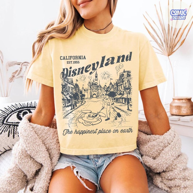 May include: Pale yellow t-shirt featuring a Disneyland graphic. The graphic shows a street scene with the Disneyland logo, the text "California Est. 1955", and the phrase "The happiest place on earth".