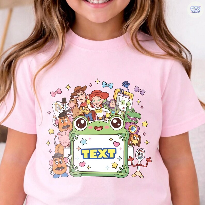 May include: A light pink t-shirt featuring a colorful graphic print of various animated characters surrounding a central green frog-shaped frame. The frame contains the word TEXT in bold blue letters, accented by small stars, hearts, and bows.
