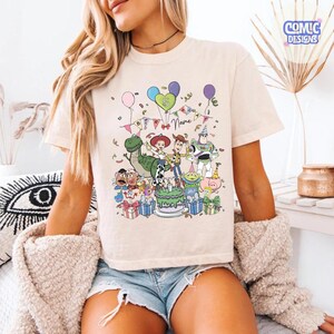 Puede incluir: Una camiseta de color crema con un estampado gr&aacute;fico de varios personajes de juguete animados, incluyendo un vaquero, un guardi&aacute;n espacial, una vaquera y un dinosaurio, rodeando un pastel de cumplea&ntilde;os con globos y regalos.
