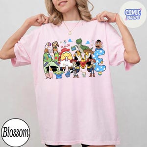 May include: A light pink t-shirt featuring a graphic print of the word NAME, with each letter filled with patterns inspired by Toy Story characters. The design includes illustrations of Woody, Buzz Lightyear, Jessie, Rex, Slinky Dog, and Mr. Potato Head.