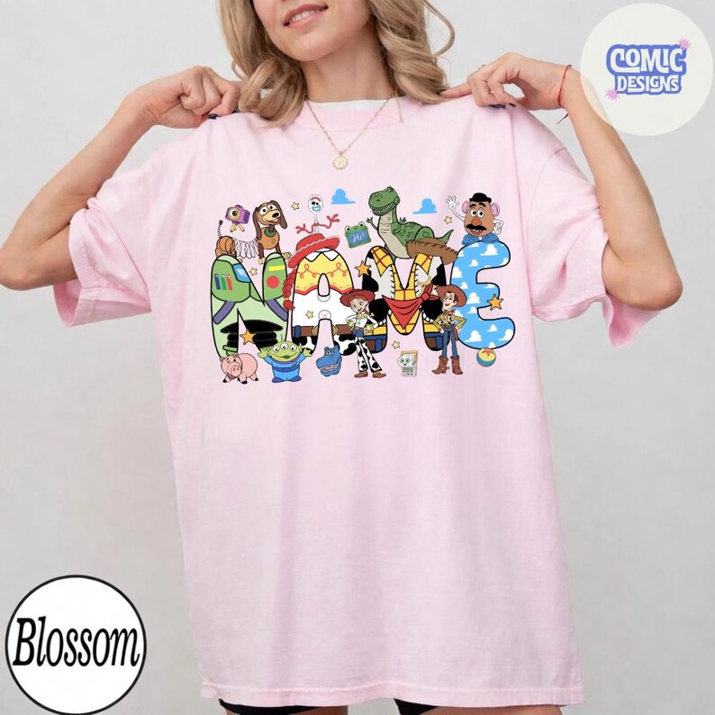 May include: A light pink t-shirt featuring a graphic print of the word NAME, with each letter filled with patterns inspired by Toy Story characters. The design includes illustrations of Woody, Buzz Lightyear, Jessie, Rex, Slinky Dog, and Mr. Potato Head.
