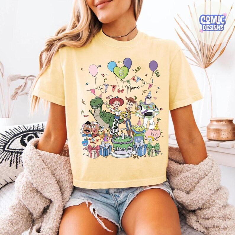 Puede incluir: Una camiseta de color amarillo p&aacute;lido con un gr&aacute;fico colorido de personajes de Toy Story, incluyendo a Woody, Buzz Lightyear, Jessie, Rex y el Sr. Patata en una fiesta de cumplea&ntilde;os con globos, regalos y un pastel. El texto dice "5" y "Name".