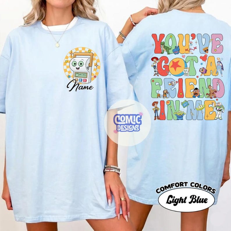 Op de afbeelding: Een lichtblauw t-shirt met een afbeelding op de borst van een stripfiguur in een geel geblokte cirkel met daaronder het woord Name. De achterkant toont kleurrijke Toy Story-illustraties en de tekst YOU'VE GOT A FRIEND IN ME.